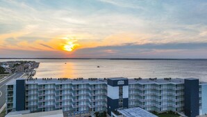 A multi-story hotel with a rooftop pool area, located near a waterfront with a sunset view. - Salty Air & Sunset Chairs | Bayfront Bliss in OCMD (Ocean City)