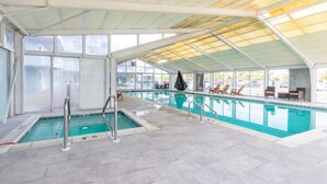 An indoor swimming pool with clear glass walls, a covered roof, and lounge chairs along the side. - Salty Air & Sunset Chairs | Bayfront Bliss in OCMD (Ocean City)