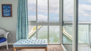 A room with a large sliding glass door leading to a view of a waterway and a bridge. There is a white chair with a blue cushion and a light blue upholstered bench. - Fries, Ferris Wheels & Bay Views | Your OCMD Getaway (Ocean City)