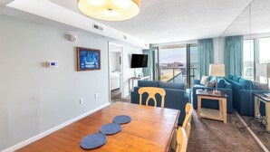 A modern hotel room with a dining area, a living space with a sofa and TV, and a large window offering a city view. - Fries, Ferris Wheels & Bay Views | Your OCMD Getaway (Ocean City)