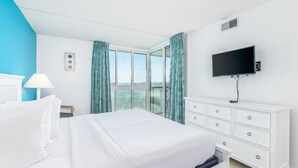 A hotel room with a large bed, a TV mounted on the wall, and a view of the outdoors through the windows. - Crab Feast Headquarters | Bay Views & Beach Days Await (Ocean City)