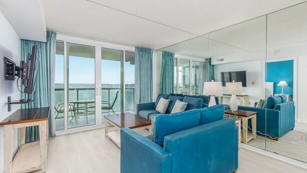 A modern living room with a large sofa, a coffee table, and a TV. There is a balcony with a table and chairs, and a large mirror on the wall. - Crab Feast Headquarters | Bay Views & Beach Days Await (Ocean City)