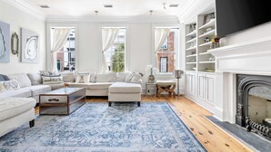 A living room with a white sofa, a coffee table, and a blue patterned rug. There is a fireplace with a mantel and a television mounted above it. - Luxury Rooftop Penthouse - King Street! (Charleston)