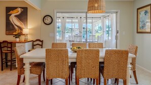 A dining area with a white table and wicker chairs, a wicker pendant light, a painting of a pelican, a clock, and a view of the outdoors through a glass door.
