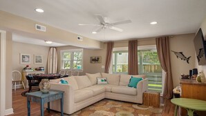 A living room with a beige sectional sofa, a small table, and a green table. There is a ceiling fan, a window with curtains, and a view of the outdoors.
