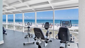Een fitnessruimte met trainingsapparatuur en grote ramen met uitzicht op een strand.