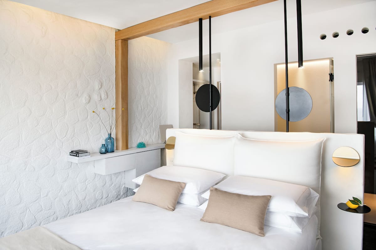 A modern bedroom with a white bed, beige pillows, a small bedside table with a vase, and a wall-mounted mirror.