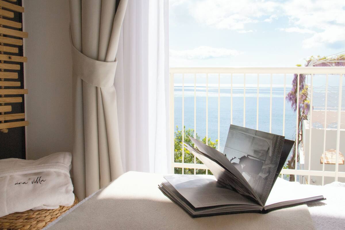 A room with a view of the sea, a bed with a white towel, and an open book on a bedside table.