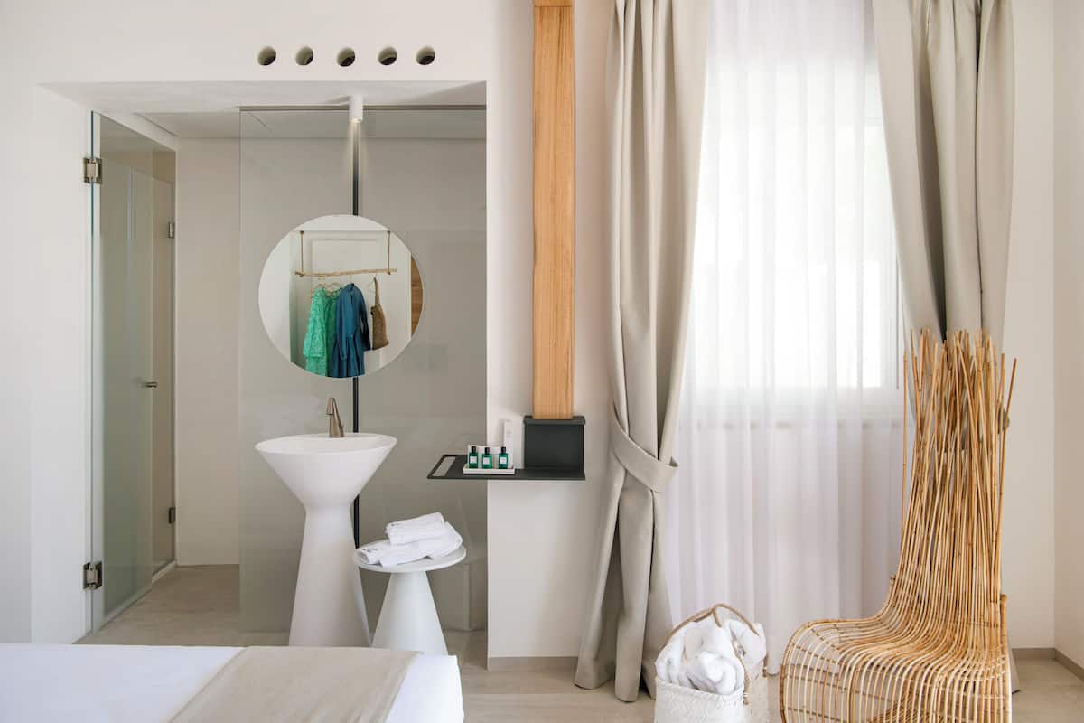 A modern hotel room with a bed, a round mirror, a sink, a small table with toiletries, and a wicker chair.