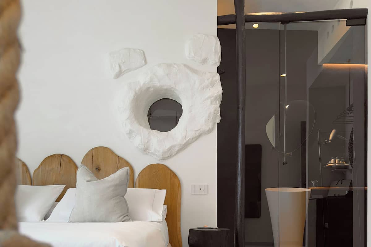 A modern bedroom with a bed, wooden headboard, a circular wall decoration, a black trash bin, and a glass partition leading to a bathroom.