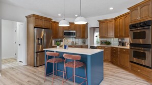 A modern kitchen with wooden cabinets, stainless steel appliances, a blue island with bar stools, and pendant lighting.