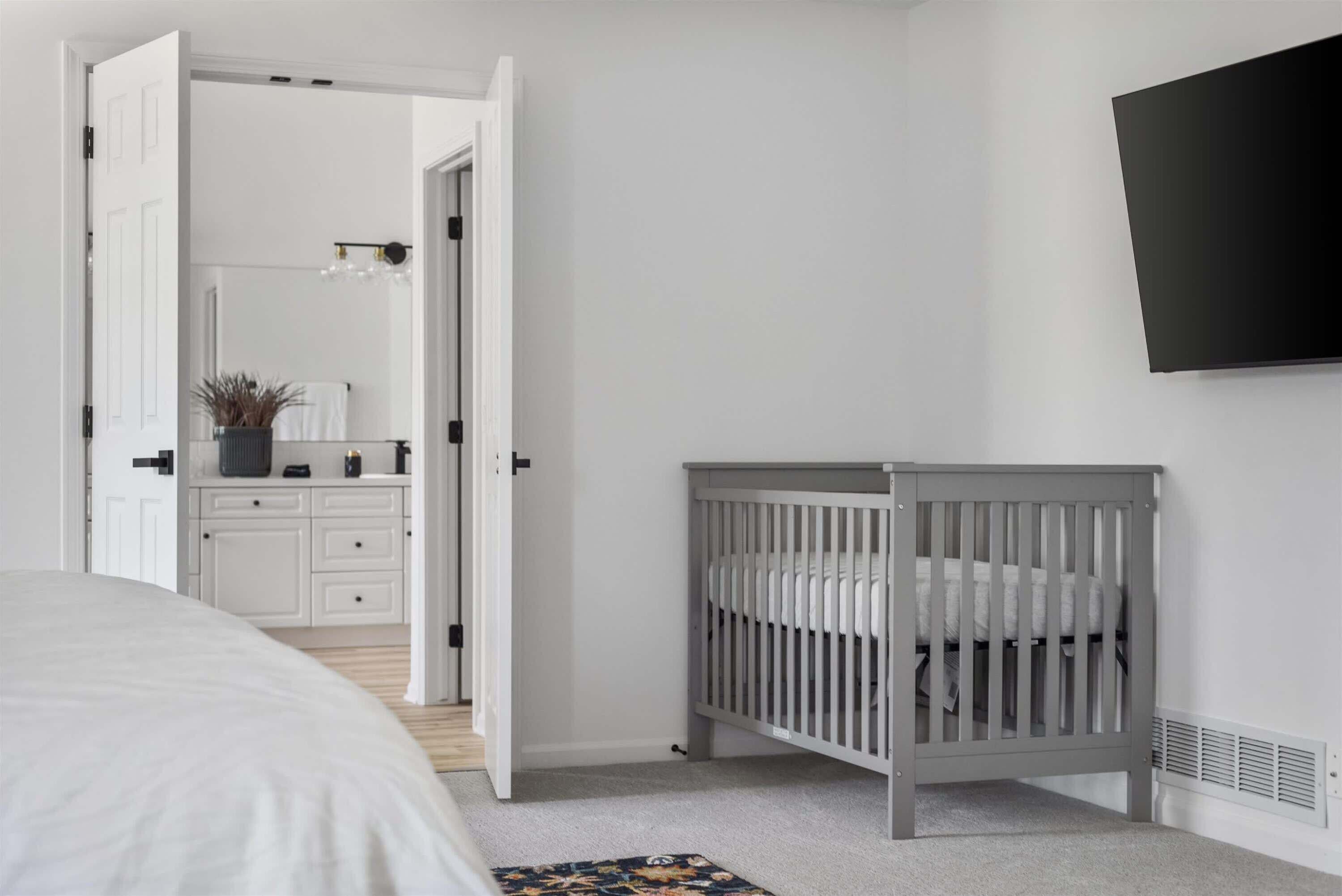Une chambre à coucher avec un lit, un berceau, un téléviseur fixé au mur et une salle de bain visible à travers une porte ouverte.