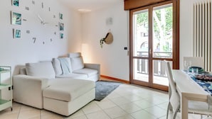 A modern living room with a white sofa, a dining table with chairs, and a large window with a view of the outdoors. - 2 Bedroom Apartment in Cesena (Cesena)