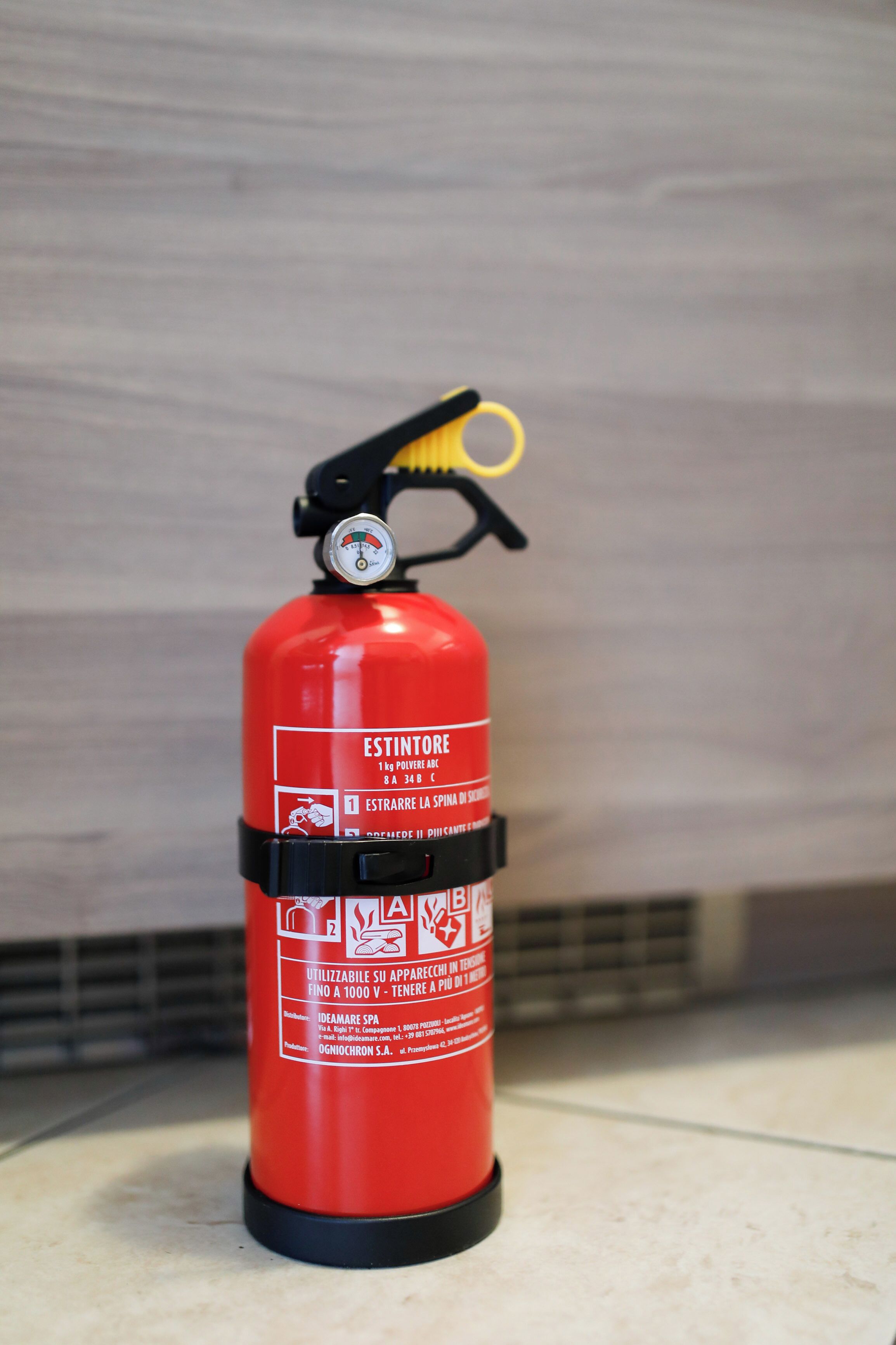 A red fire extinguisher with labels in multiple languages, mounted on a tiled floor against a wall.