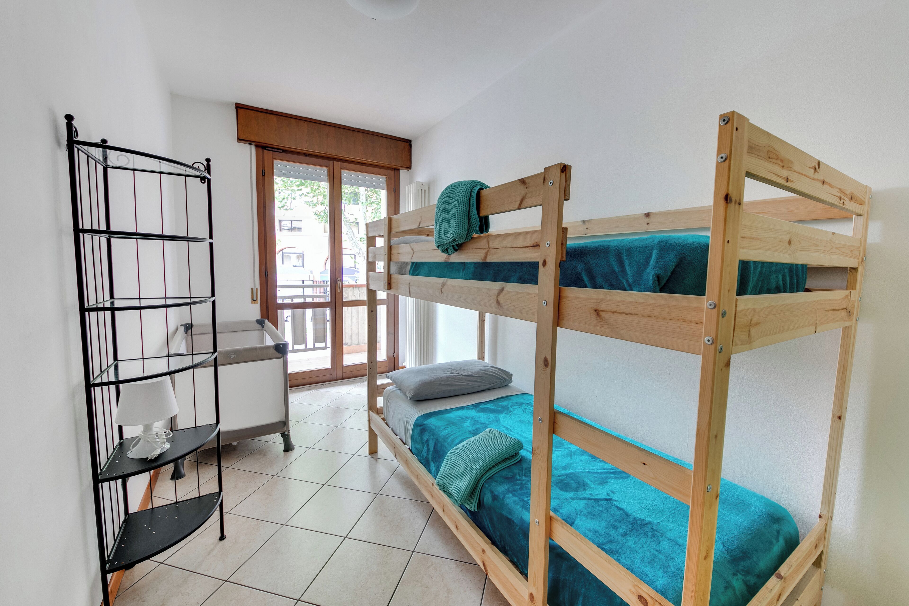 Un lit superposé avec une structure en bois et une literie de couleur turquoise, une étagère en métal avec une lampe, et une chaise blanche dans une pièce dont le sol est carrelé et qui possède une porte donnant sur l’extérieur.