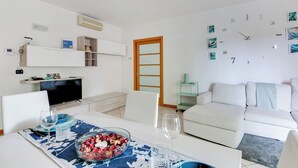 A modern living room with a white sofa, a dining table with a floral table runner, and a TV on a stand. - 2 Bedroom Apartment in Cesena (Cesena)