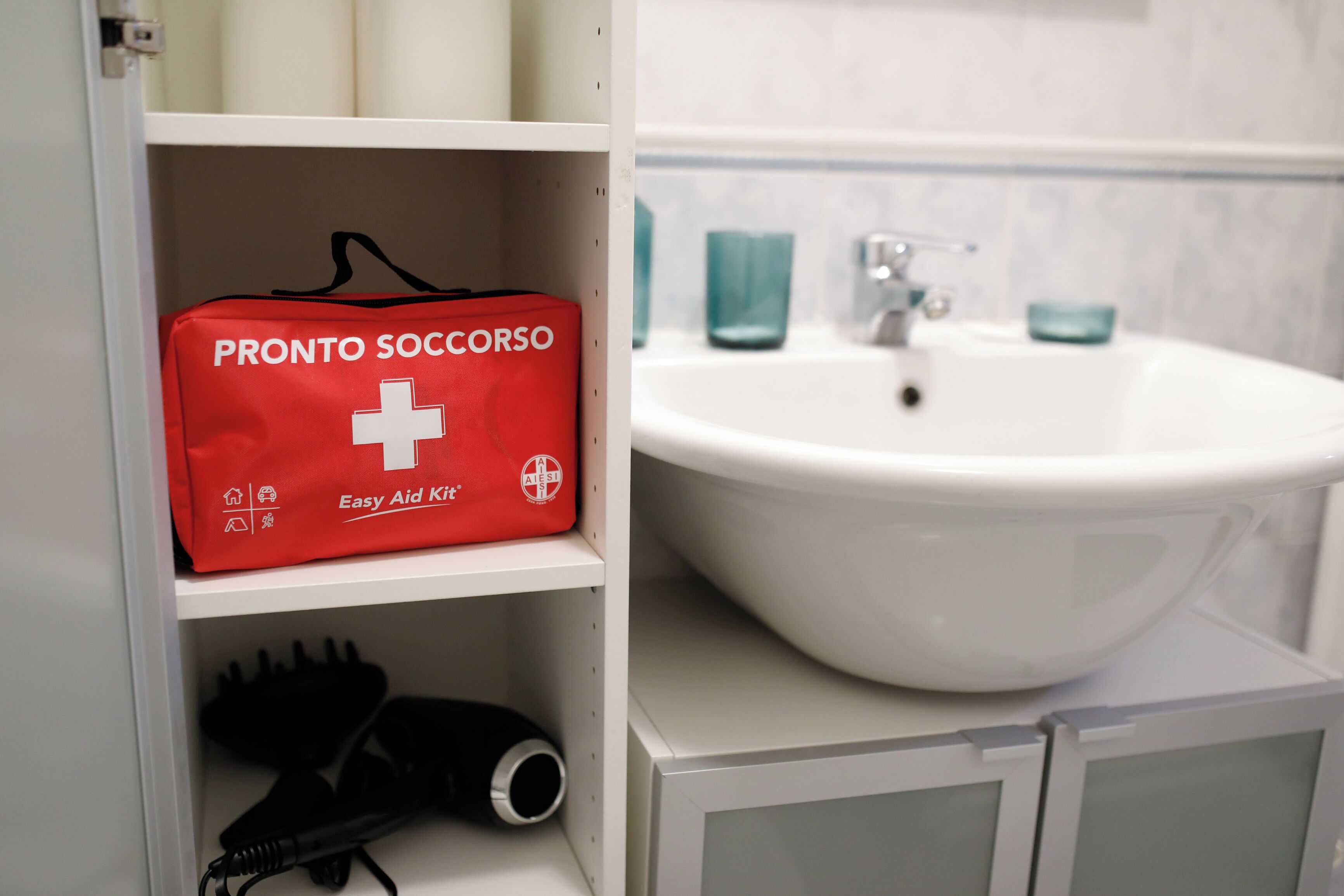 Ein Badezimmer mit einem weißen Waschbecken, einer roten Erste-Hilfe-Ausrüstung mit dem Aufdruck "PRONTO SOCCORSO" und einem Fön auf dem Regal.