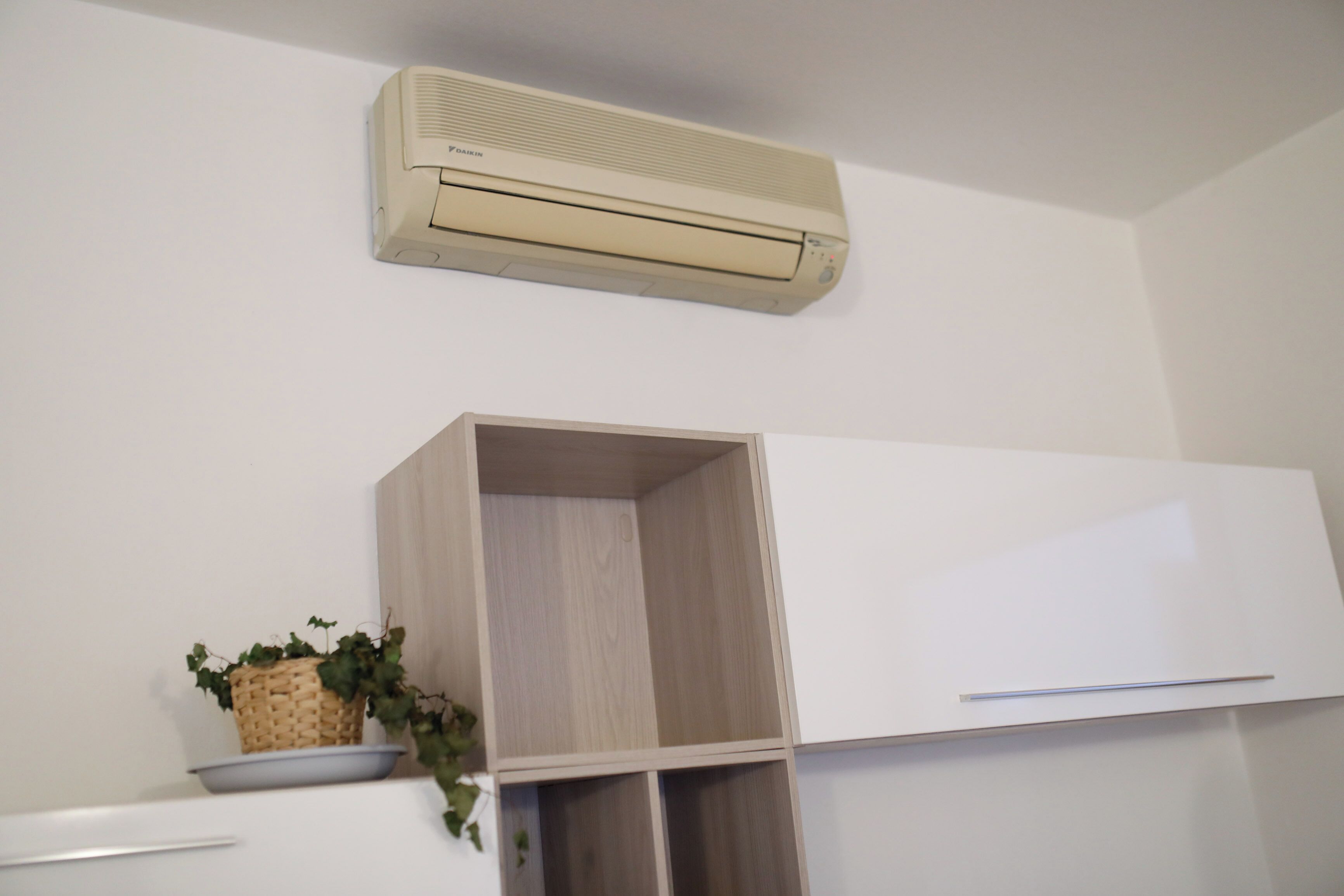 A white wall with a beige air conditioner, a wooden cabinet, and a wicker basket with green plants.