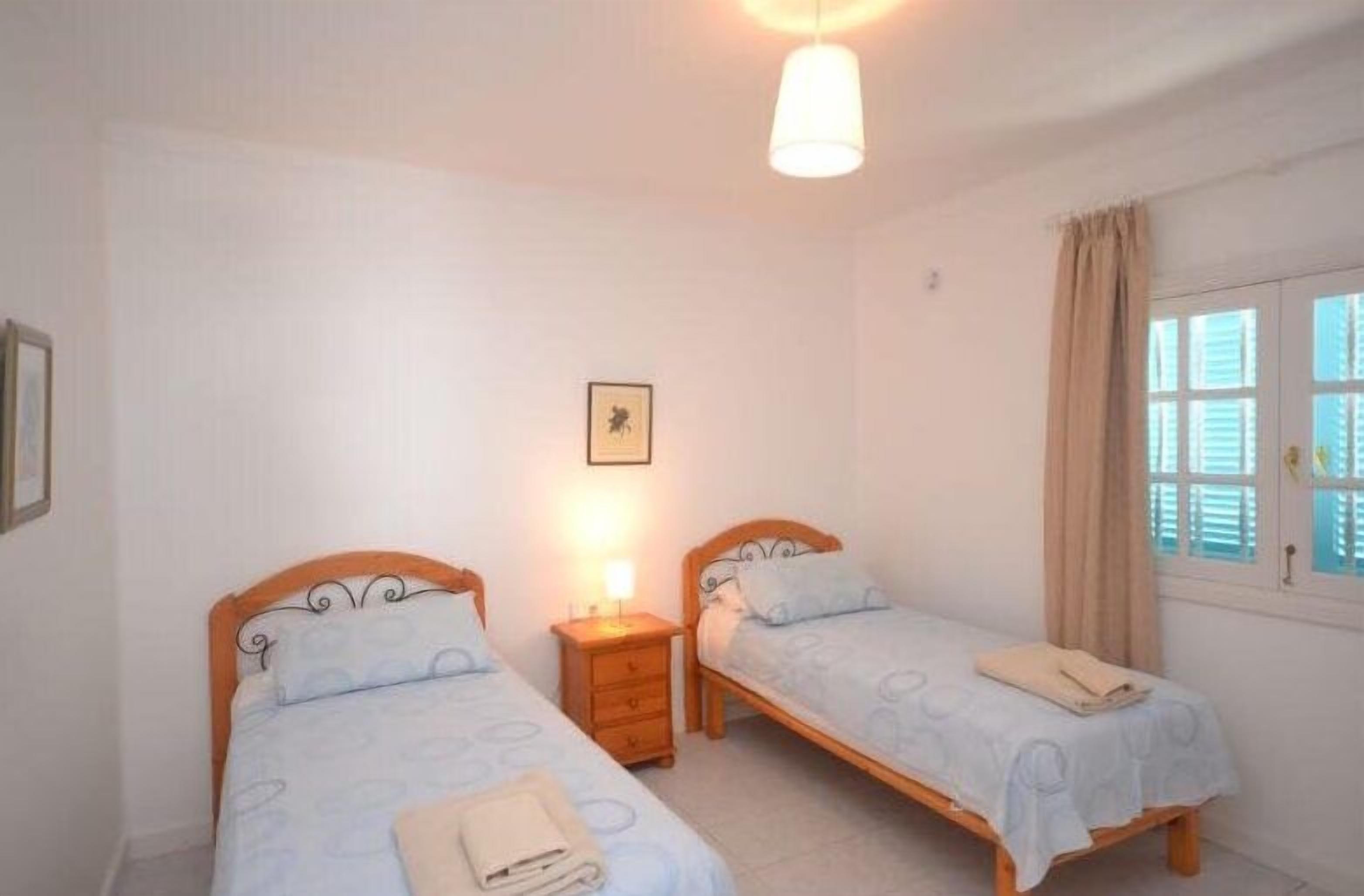 A bedroom with two single beds, a bedside table with a lamp, a framed picture on the wall, and a window with curtains.