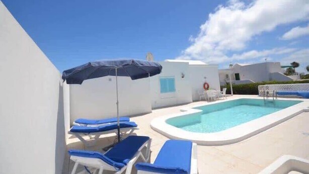 A swimming pool with blue lounge chairs and umbrellas, surrounded by white buildings. - Villa Violeta with electrically heated pool (Puerto del Carmen)
