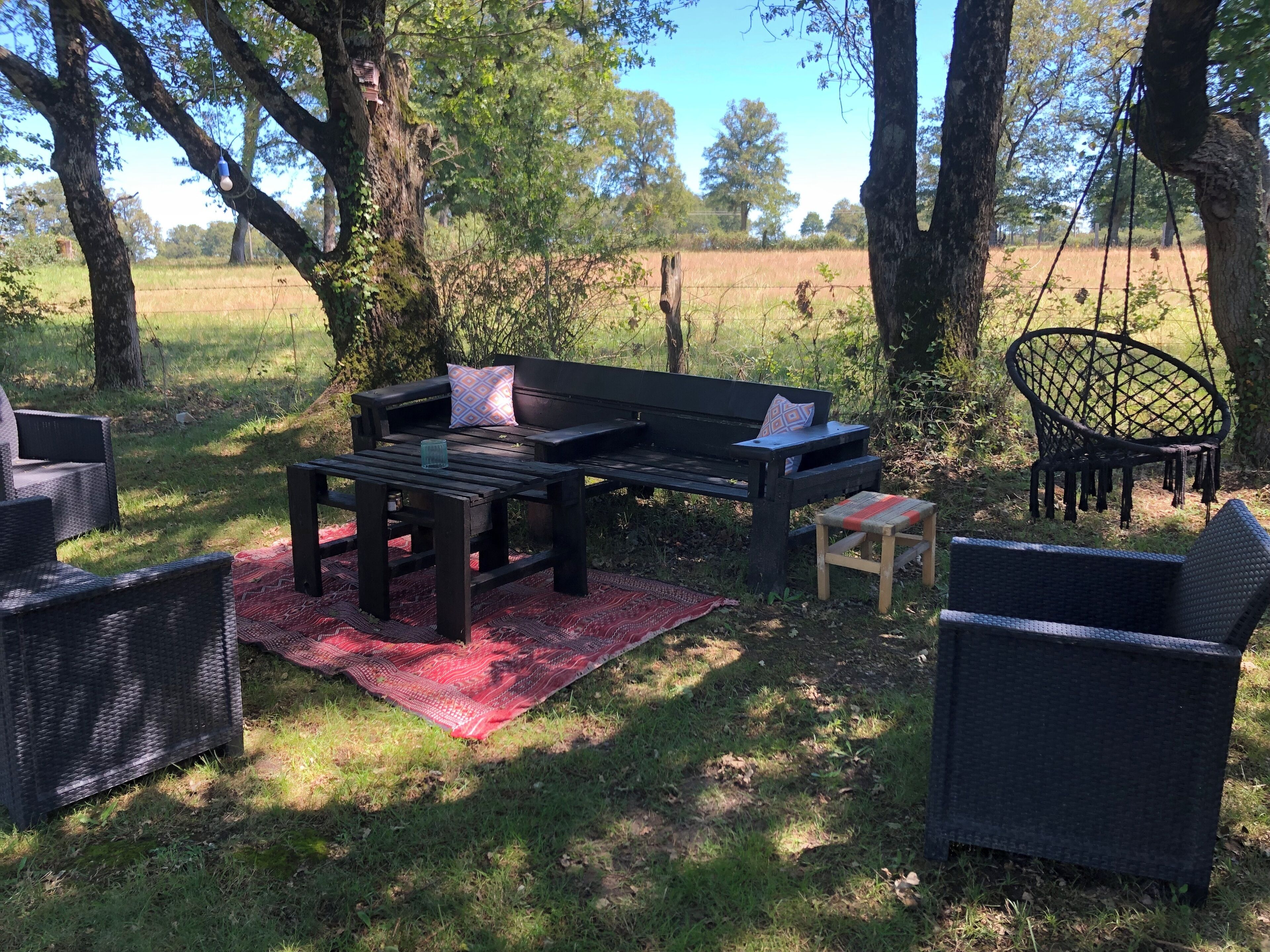 A wooden bench with cushions, a small stool, and a wicker chair in a grassy outdoor area with trees.