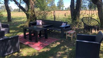 A wooden bench with cushions, a small stool, and a wicker chair in a grassy outdoor area with trees.