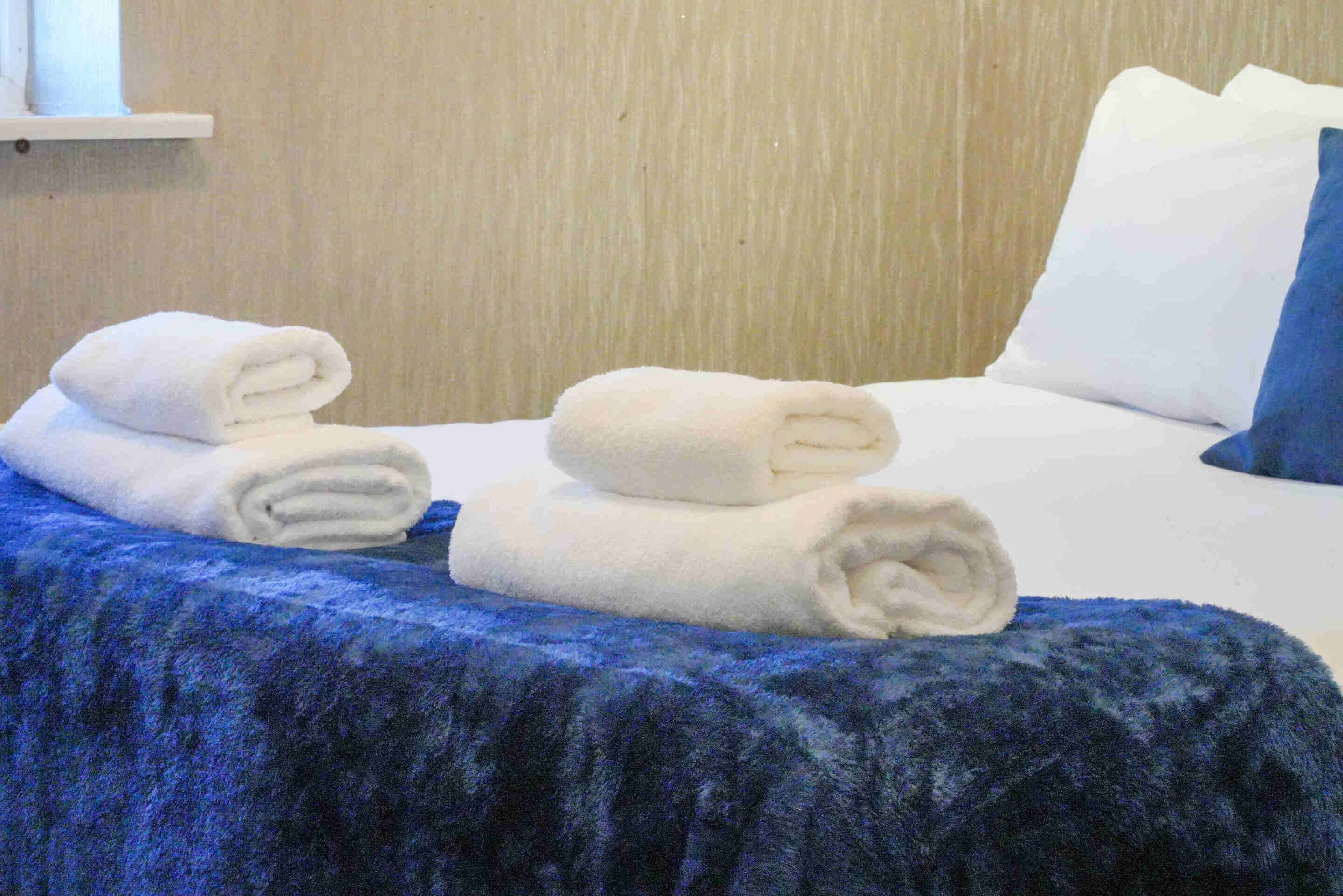 Ein Bett mit blauer Bettdecke, weißen Kissen und aufgerollten weißen Handtüchern.