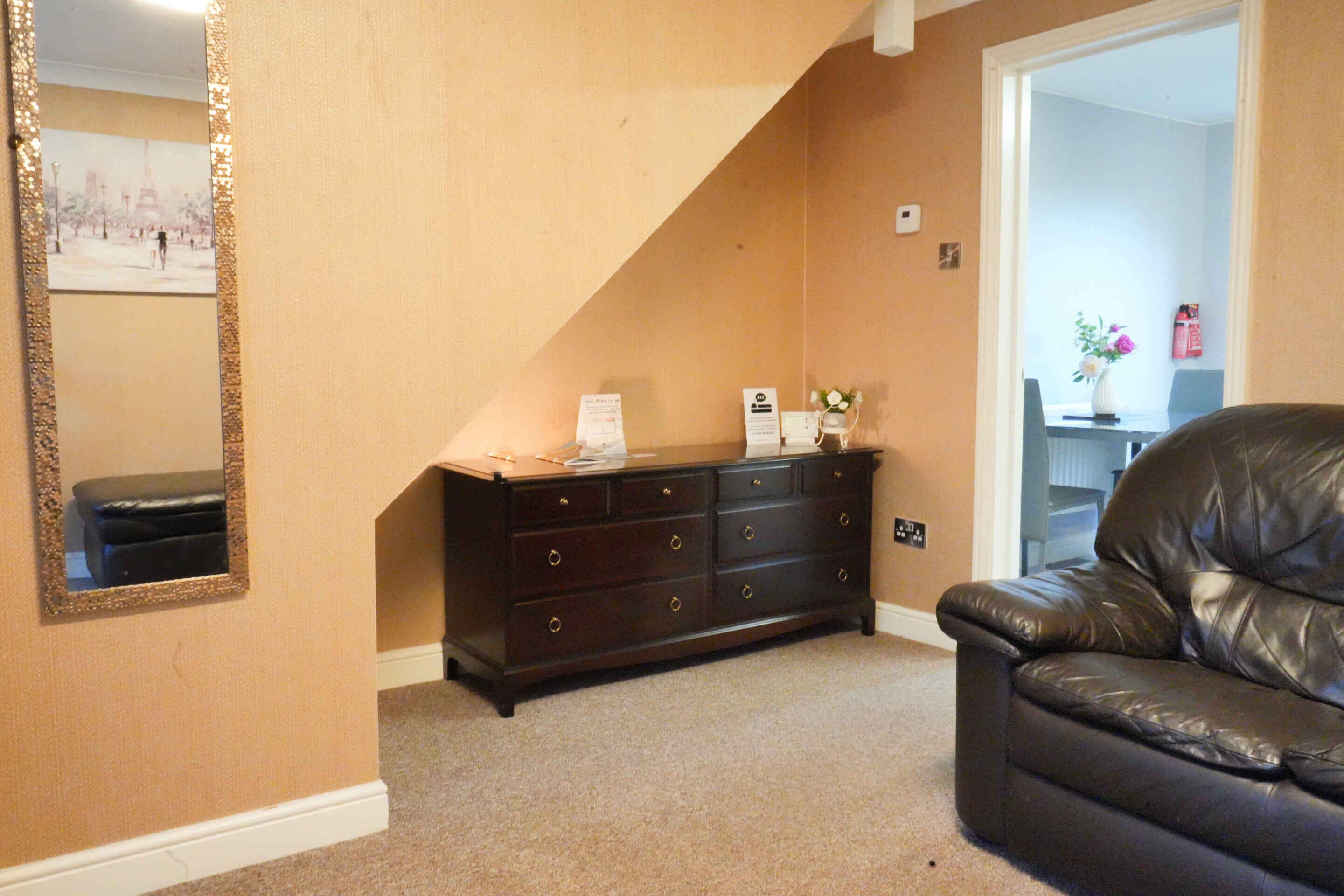 A room with a dark wooden dresser, a black leather chair, a mirror with a gold frame, and a door leading to another room.