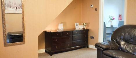A room with a dark wooden dresser, a black leather chair, a mirror with a gold frame, and a door leading to another room.