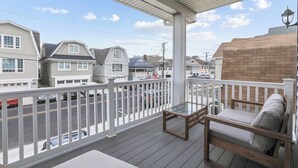 Een balkon met een glazen tafel en een beklede bank, met uitzicht op een straat met huizen en geparkeerde auto's.