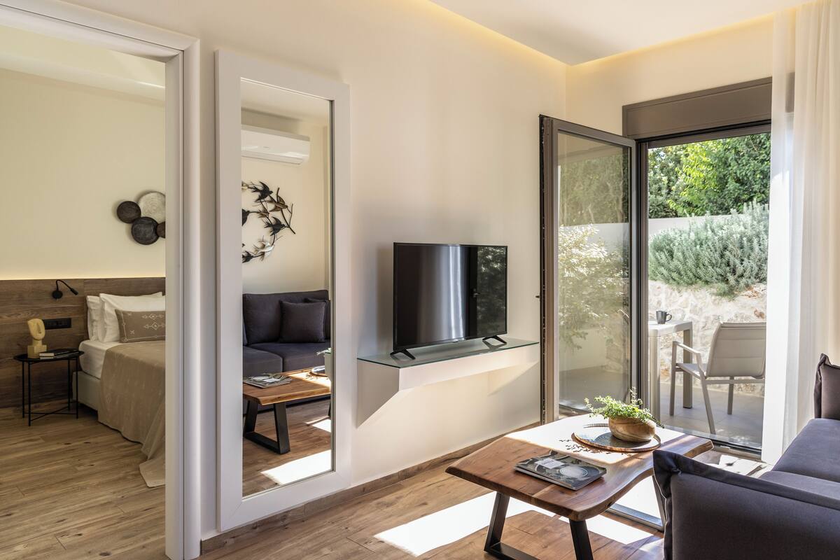 Una sala de estar moderna con sofá, una mesa de centro y televisor en un estante de vidrio. Hay una puerta corrediza de vidrio que da a un área exterior con una mesa y sillas.