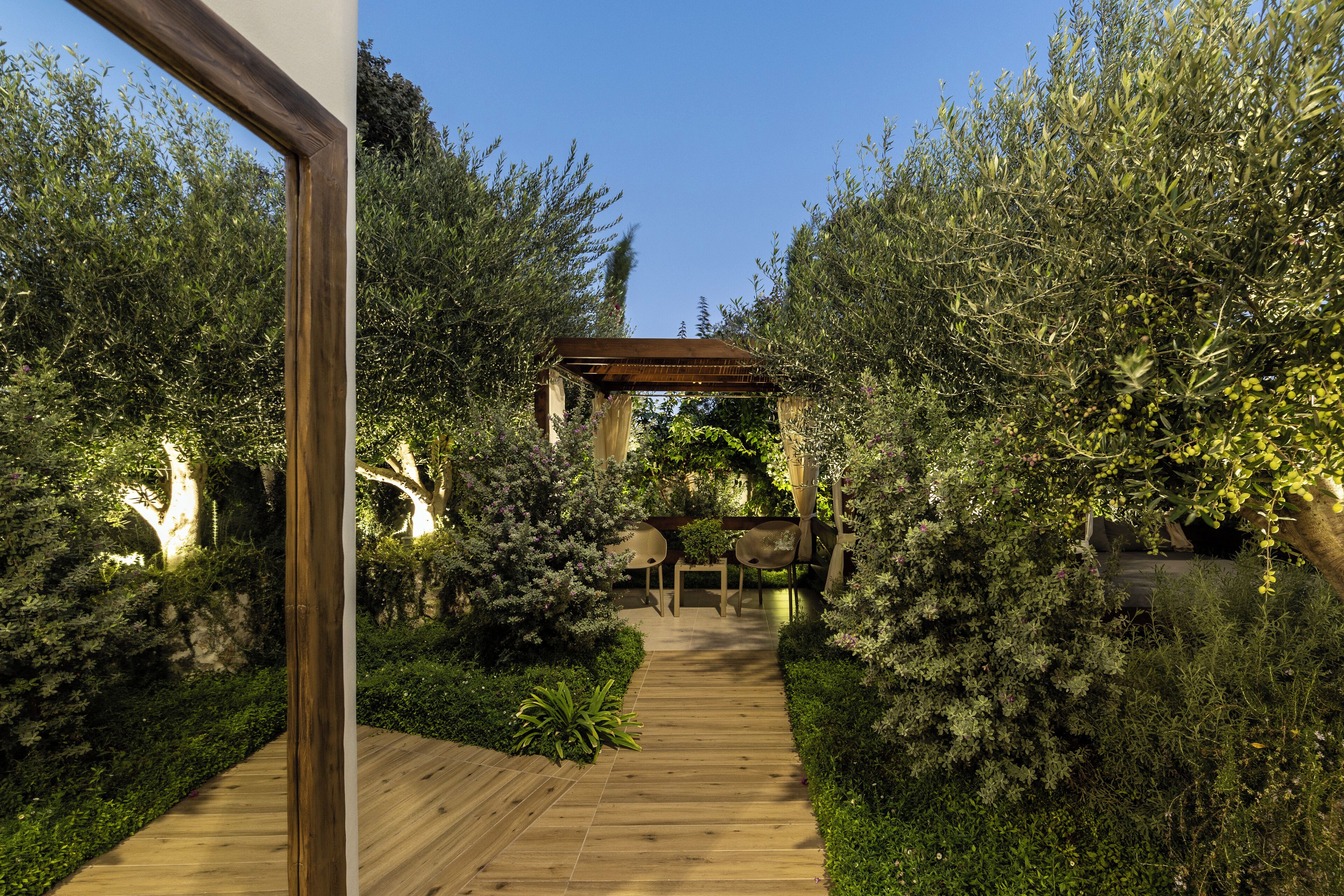 A wooden pathway leading to a gazebo in a lush garden with dense foliage and a clear sky.