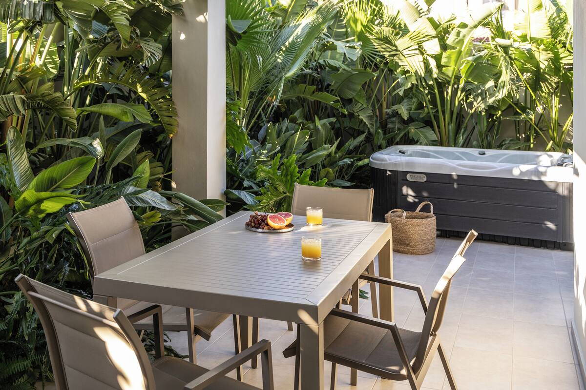 Un patio con mesa, sillas y un jacuzzi rodeado de abundante vegetación.