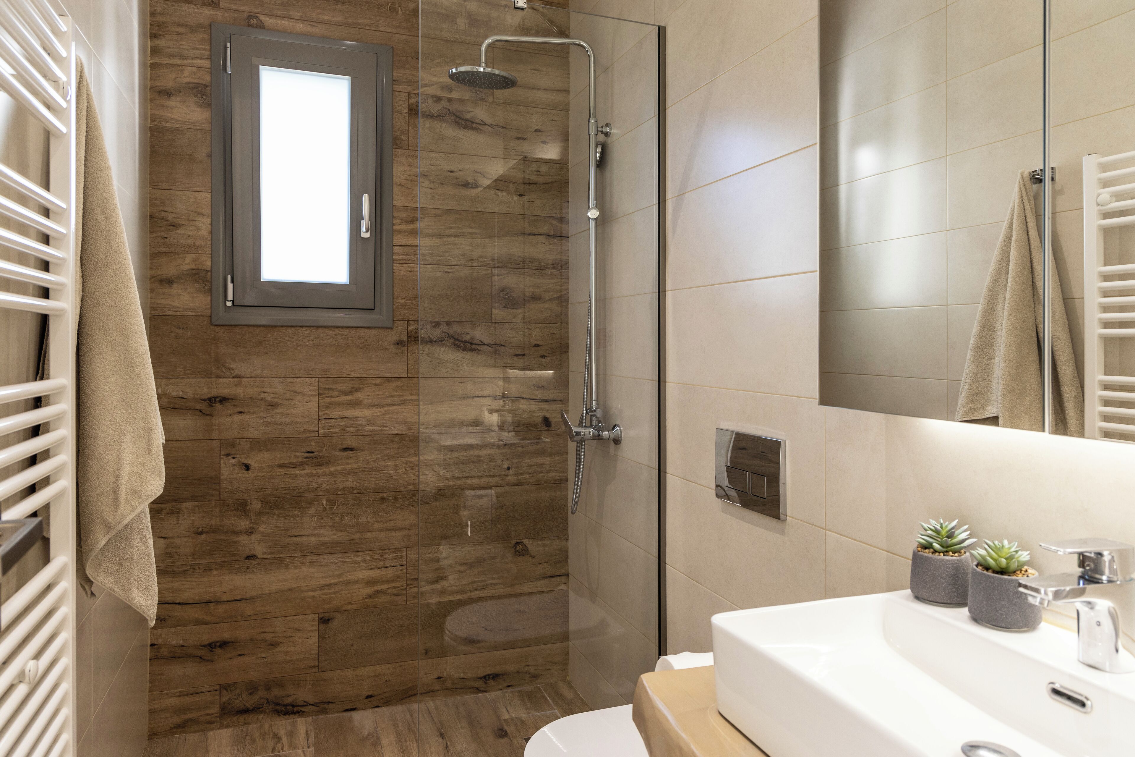 A modern bathroom with a wooden shower enclosure, a white sink with two potted plants, and a towel rack.