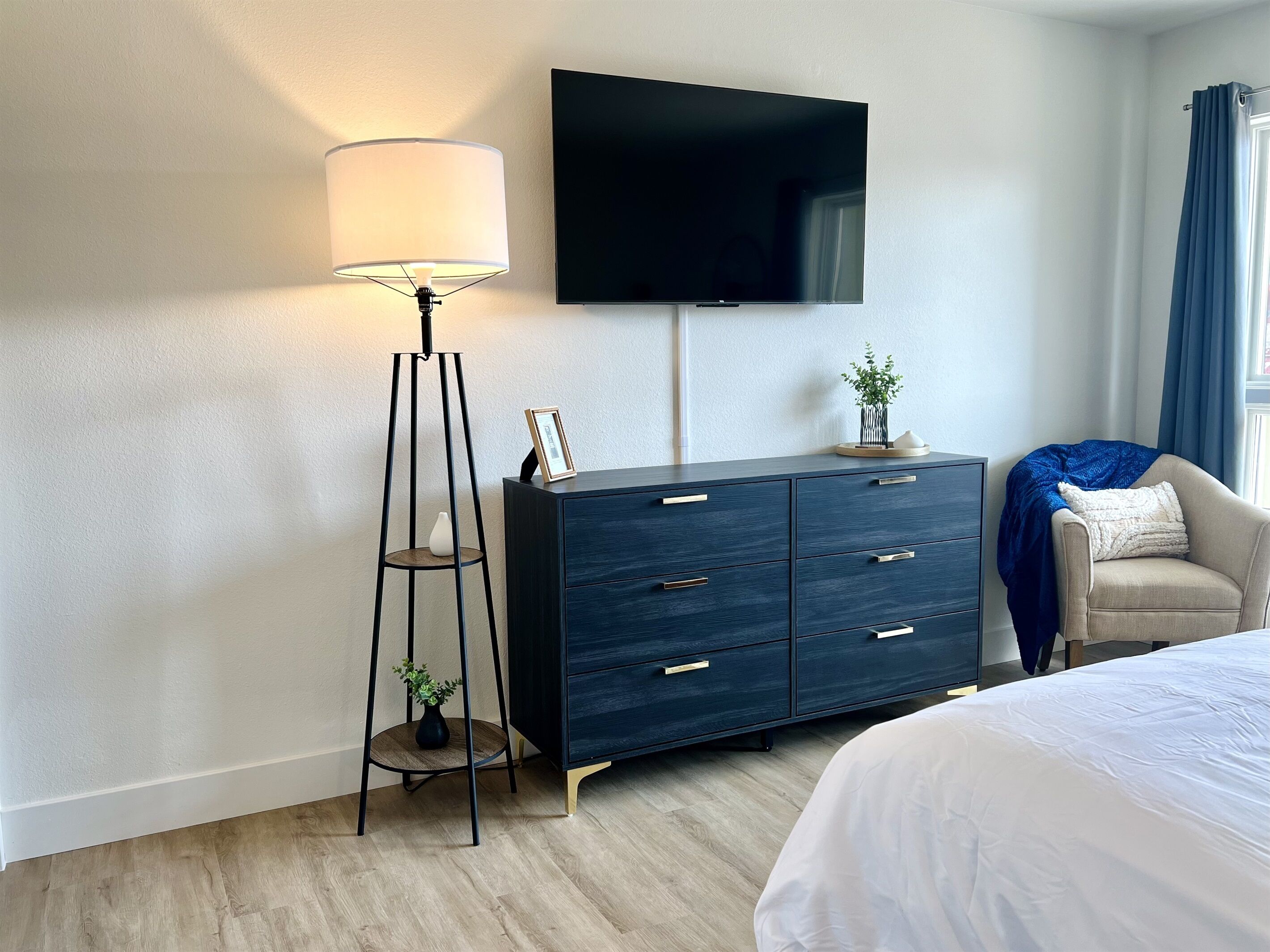 A modern hotel room with a dark wooden dresser, a standing floor lamp, a flat-screen TV mounted on the wall, a beige armchair with a blue blanket, and a large bed with white bedding.