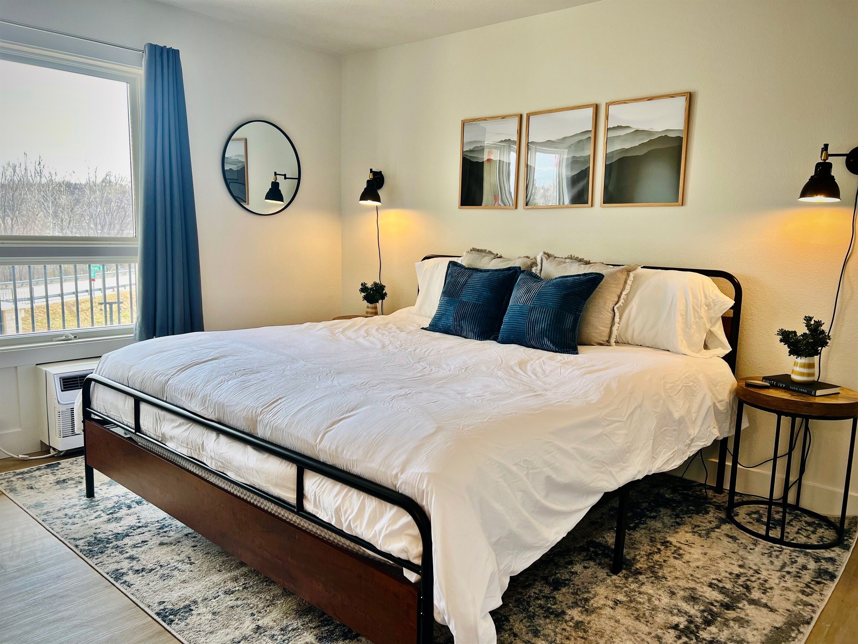 A neatly made bed with white bedding, a black metal frame, and a blue and white patterned rug. There are three wall-mounted lamps and a mirror on the wall.
