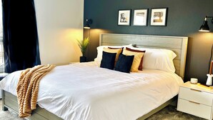 Una cama bien tendida con sábanas blancas y una manta color beige, una mesita de noche con lámpara, y un cuadro colgado en la pared.
