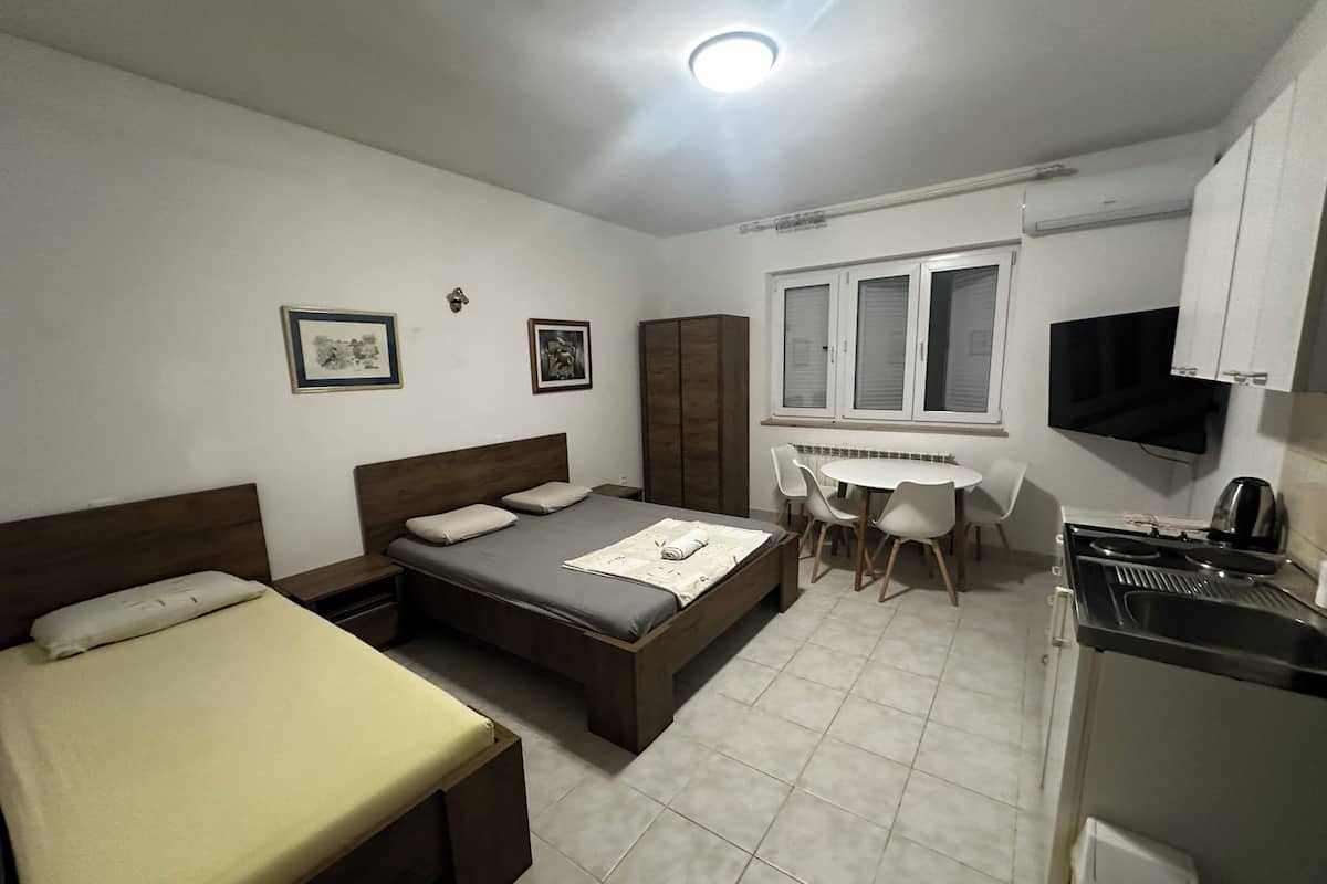 A bedroom with a bed, a bedside table, a desk with chairs, a TV, and a kitchenette.