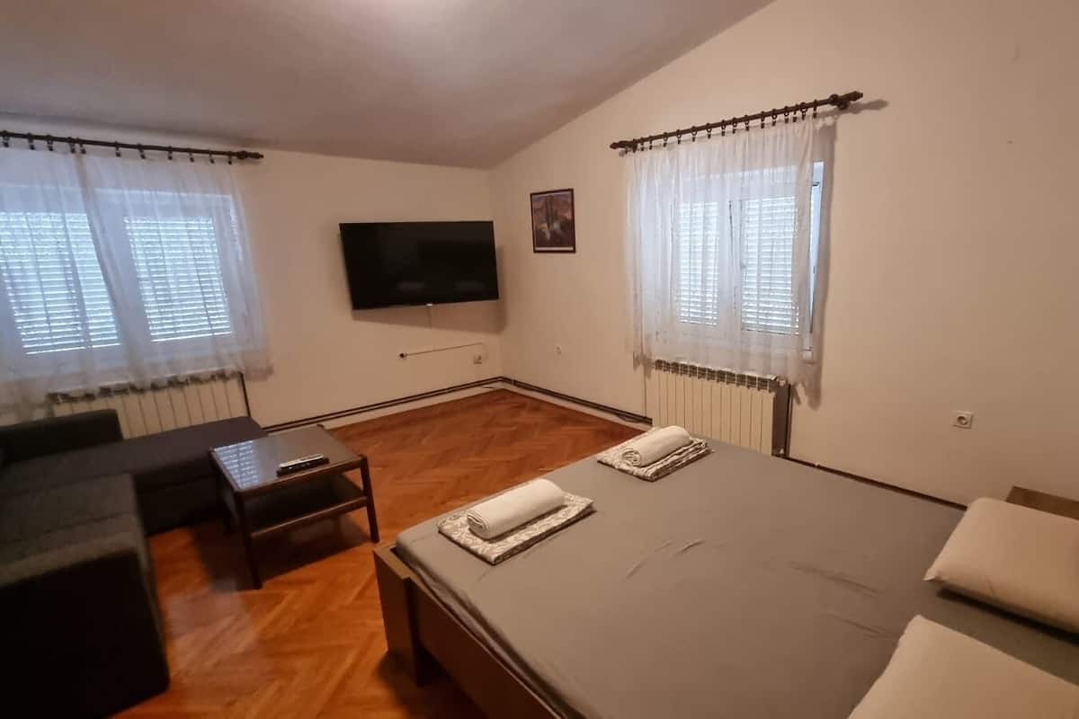 A hotel room with a bed, a small table, a sofa, a TV, and two windows with curtains.
