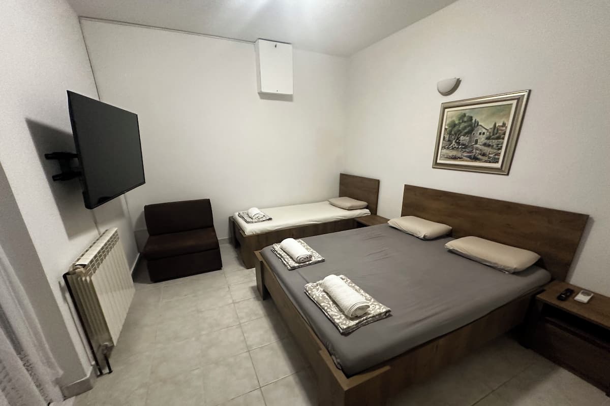 A bedroom with a large bed, two smaller beds, a chair, a TV, a radiator, and a framed picture on the wall.