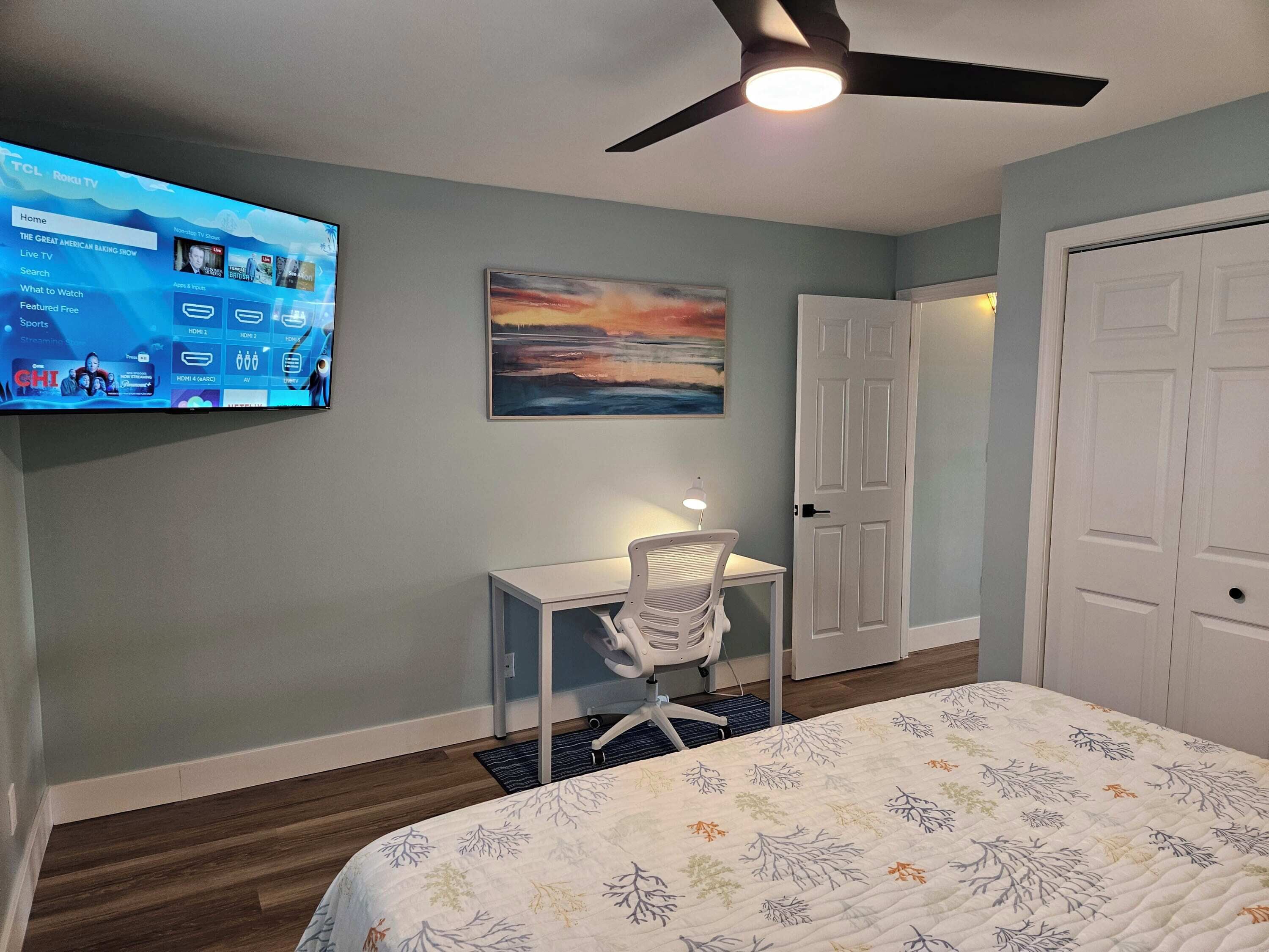 A bedroom with a bed, a desk, a chair, a painting, and a TV displaying a Roku TV interface.