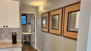 Eine moderne Küche mit weißen Schränken, einer Arbeitsplatte aus Granit und einer Kaffeemaschine. An der Wand befinden sich spiegel mit Holzrahmen und im Hintergrund ein Fernseher.