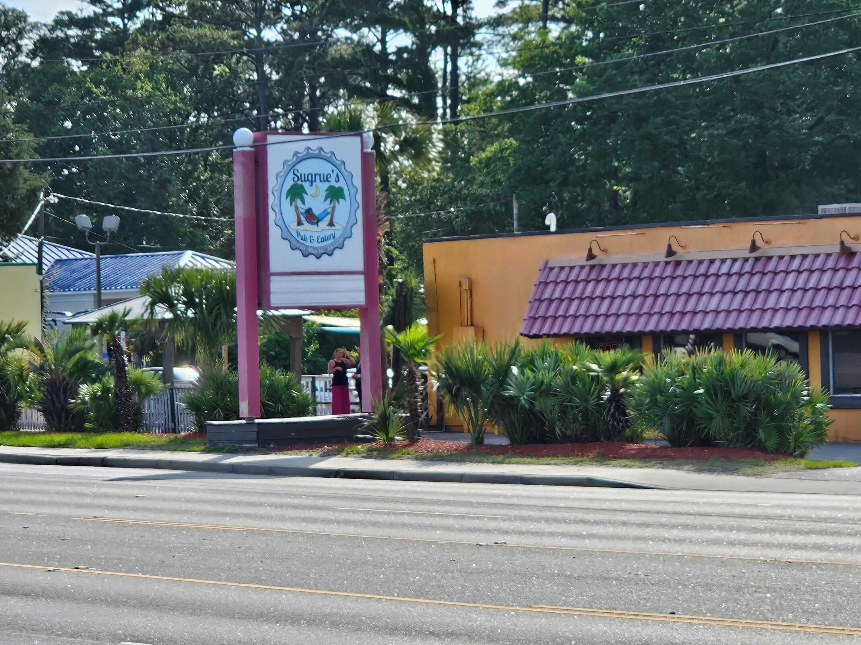 A sign for Sugrue's Pub & Cafe with a tropical logo, surrounded by greenery and a building with a purple roof.