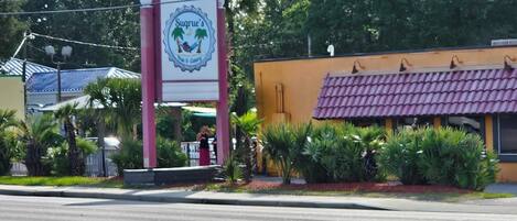 A sign for Sugrue's Pub & Cafe with a tropical logo, surrounded by greenery and a building with a purple roof.