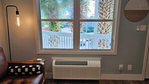 Ein Zimmer mit einem braunen Ledersofa, einer Stehlampe, einem Fenster mit Blick auf einen Balkon und Palmen sowie einer Wand-Klimaanlage.