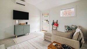 Een woonkamer met een flatscreen televisie, een groen kastje, een beige bank en een raam met uitzicht op bomen.