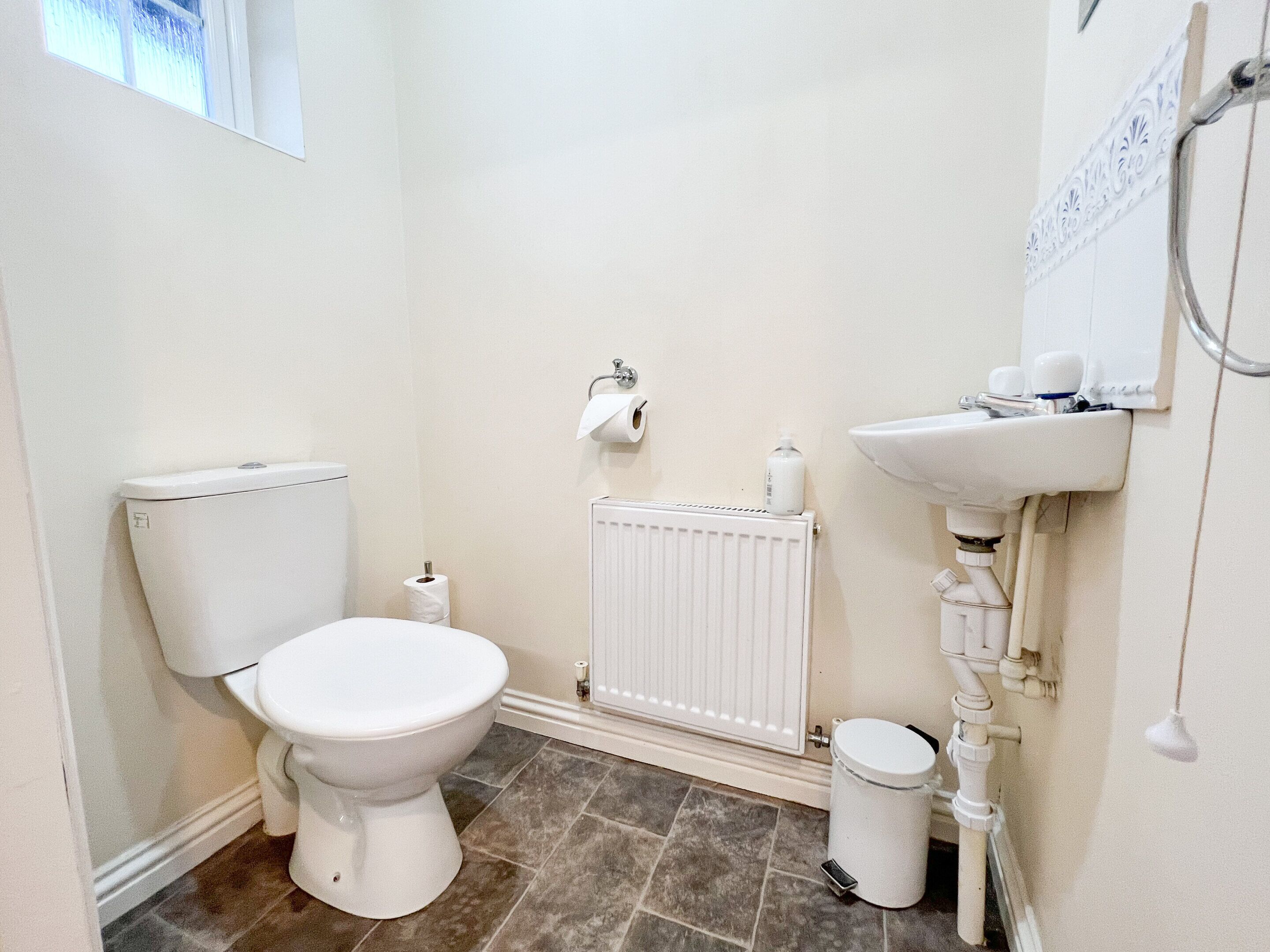 A small, white bathroom with a toilet, a radiator, a sink, and a trash bin.