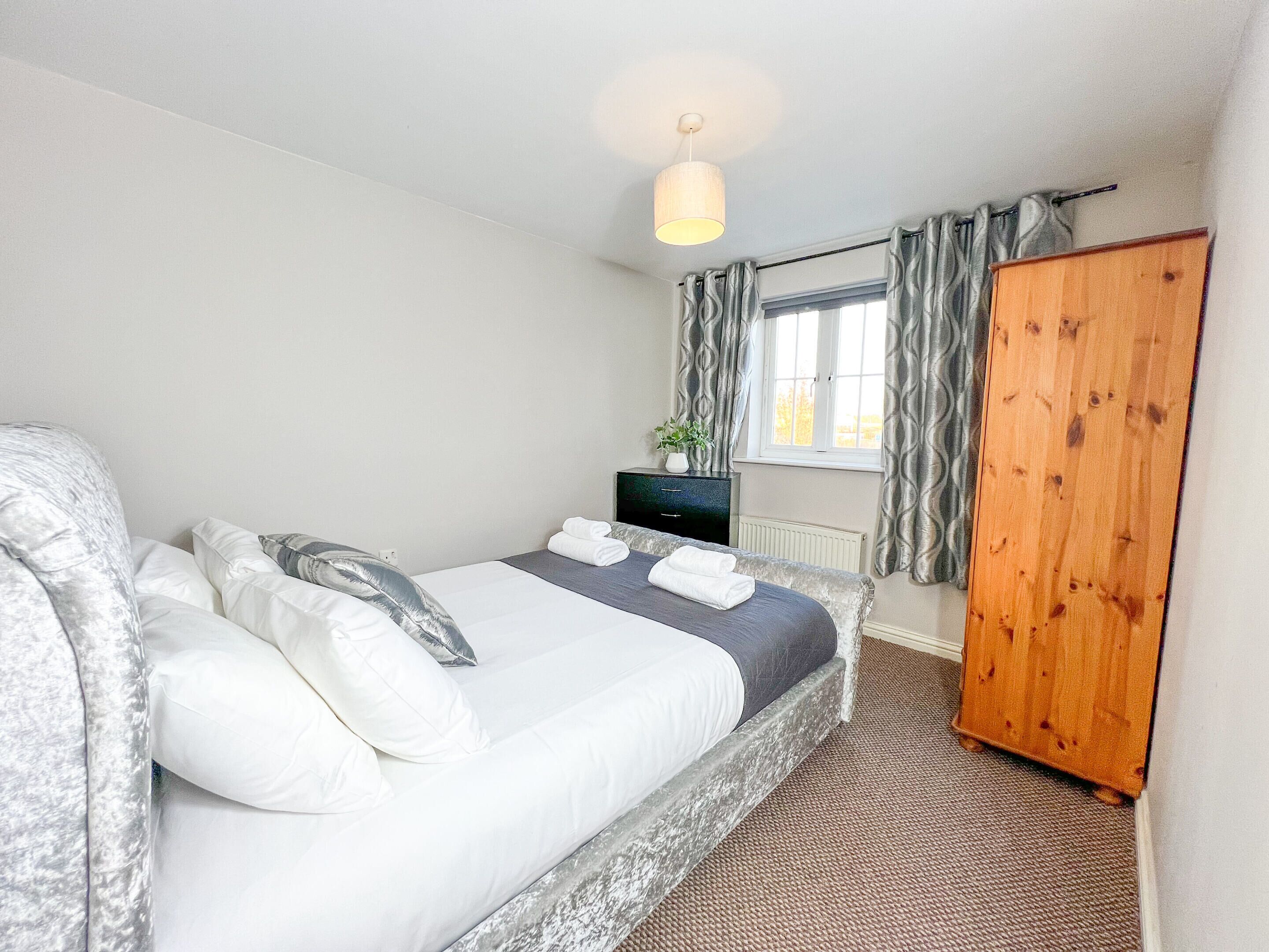 A bedroom with a bed, a wooden wardrobe, a dresser, a window with curtains, and a hanging light fixture.