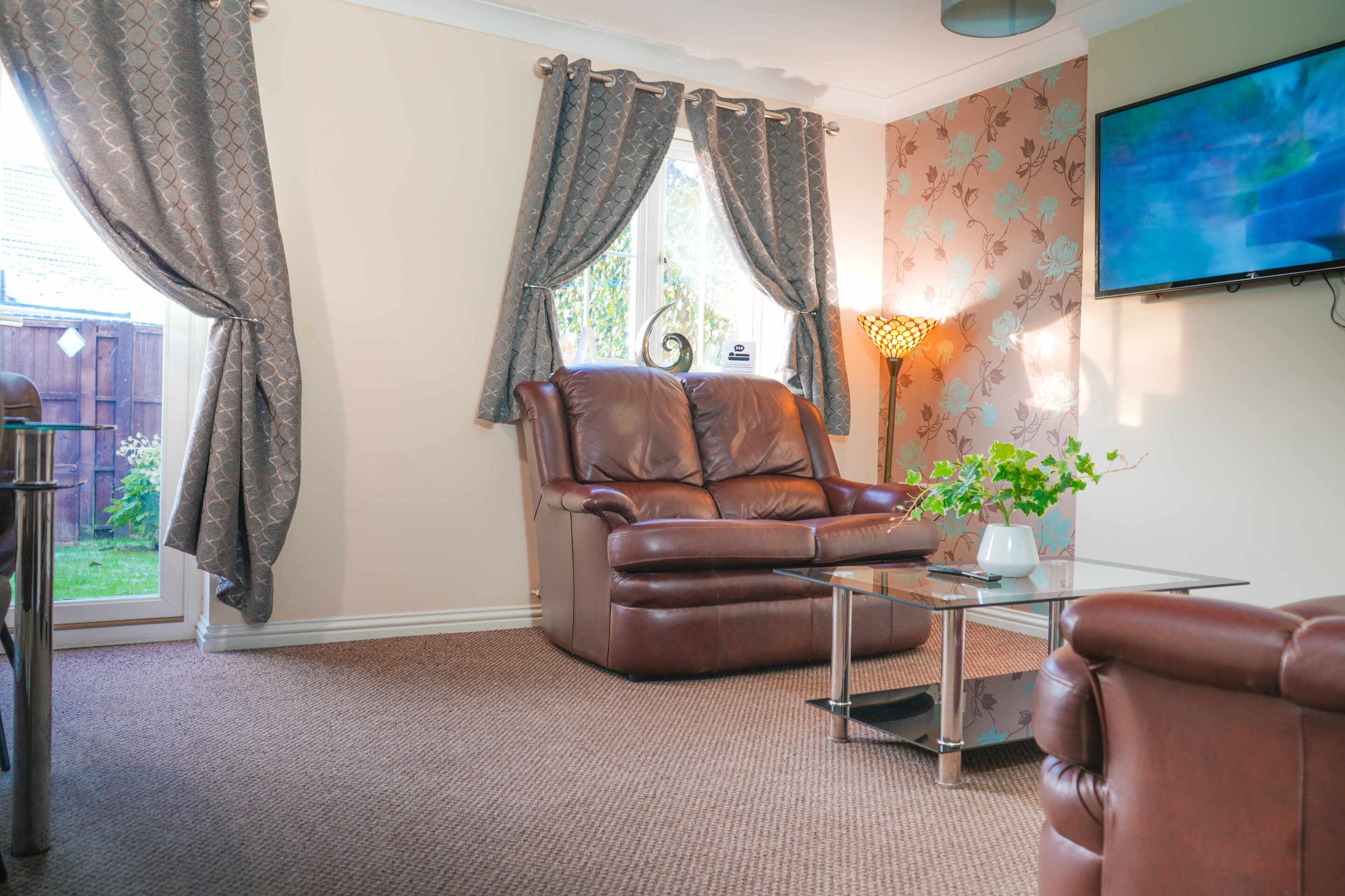 A living room with a leather sofa, a glass coffee table, a TV mounted on the wall, and a window with curtains.
