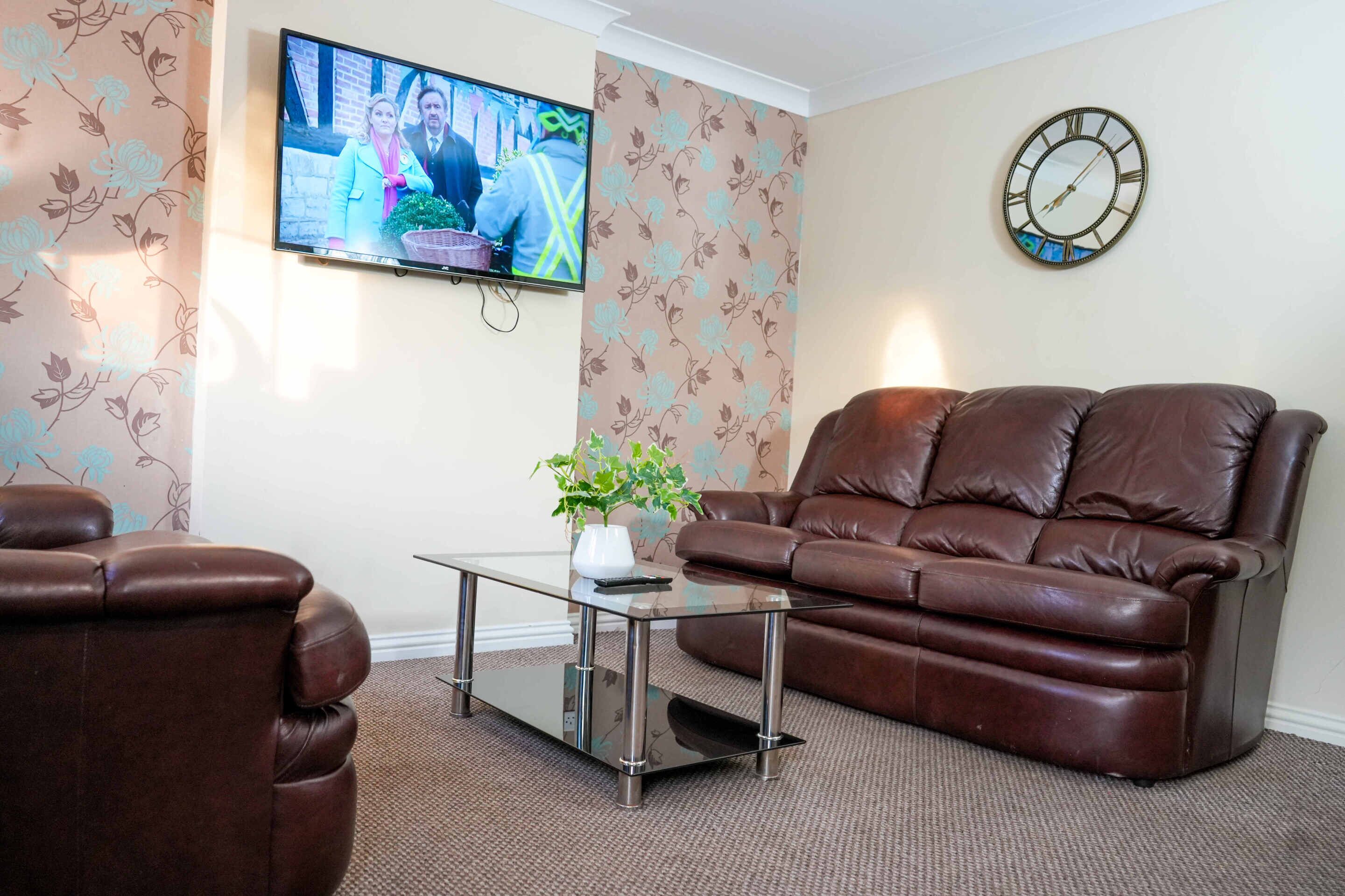 A living room with a brown leather sofa, a glass coffee table, a wall-mounted TV, and a clock.